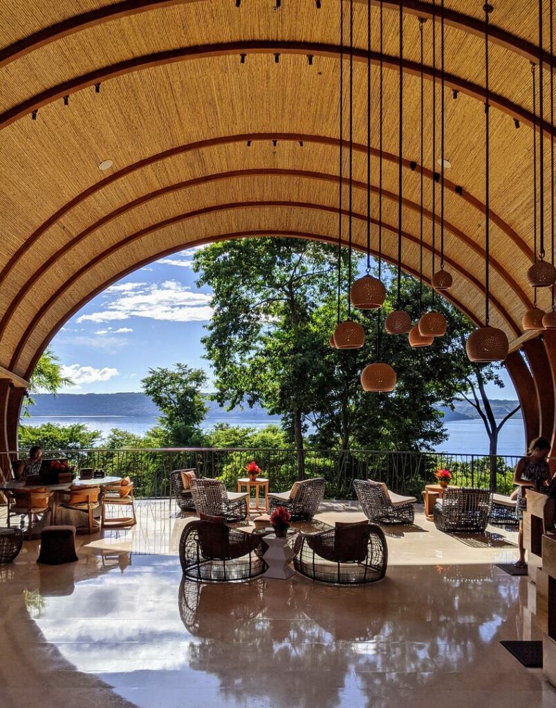 A spacious lounge with curved wooden arches and hanging lamps, featuring wicker chairs and tables. Large windows offer a view of lush trees and the ocean under a bright blue sky. Sunlight reflects on the polished floor.