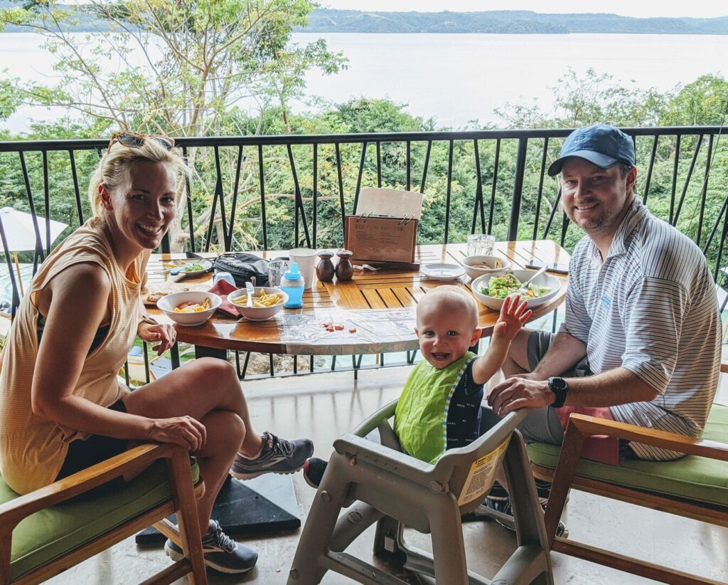 A smiling family sits at an outdoor restaurant table overlooking a scenic landscape with water and greenery. A woman, a toddler in a high chair, and a man enjoy the meal. The toddler waves at the camera. Plates of food are on the table.