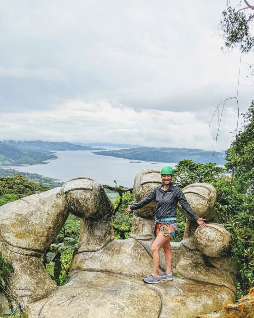 A person in outdoor gear and a green helmet stands on a giant stone hand sculpture in a lush landscape. A lake and mountains are visible in the background under a cloudy sky.