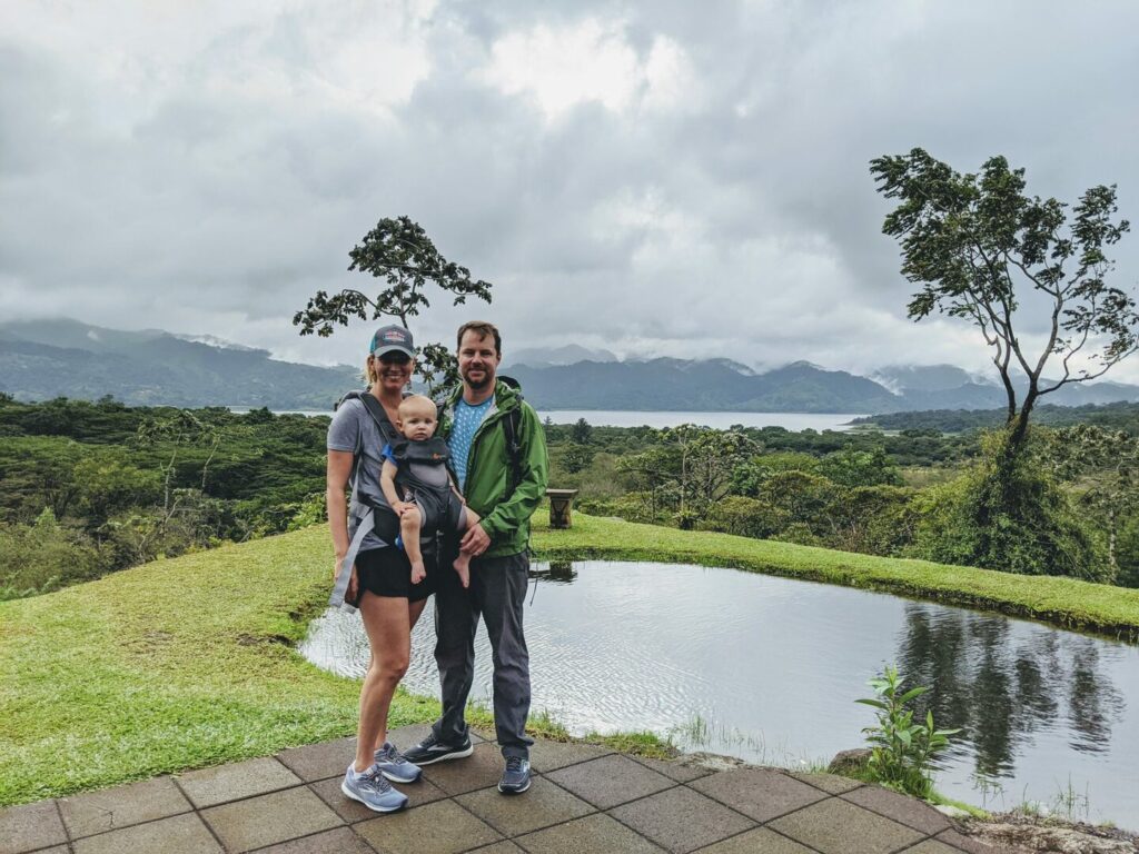 A family of three stands smiling in front of a pond with lush greenery and mountains in the background. The woman holds a baby, and both adults are wearing casual outdoor clothing. The sky is overcast, hinting at a recent or upcoming rain.