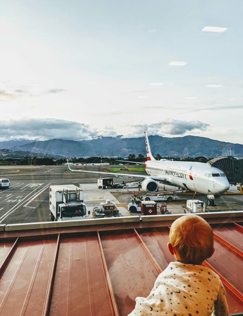 A toddler with blonde hair, wearing a white patterned jacket, looks out of an airport window at a parked airplane. The scene includes baggage carts and a mountainous landscape with a cloudy sky in the background.