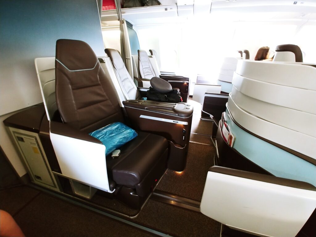 Spacious airplane business class seats with leather upholstery, armrests, and adjustable headrests. One seat has a blue blanket on it. Bright natural light enters from windows in the background.