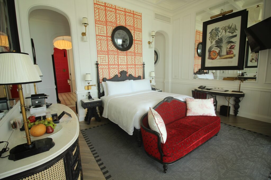 Elegant hotel room with a white bed, red-patterned sofa, fruit bowl on a table, wall mirrors, and decorative wallpaper. The room features classic furniture and a large botanical artwork above the bed.