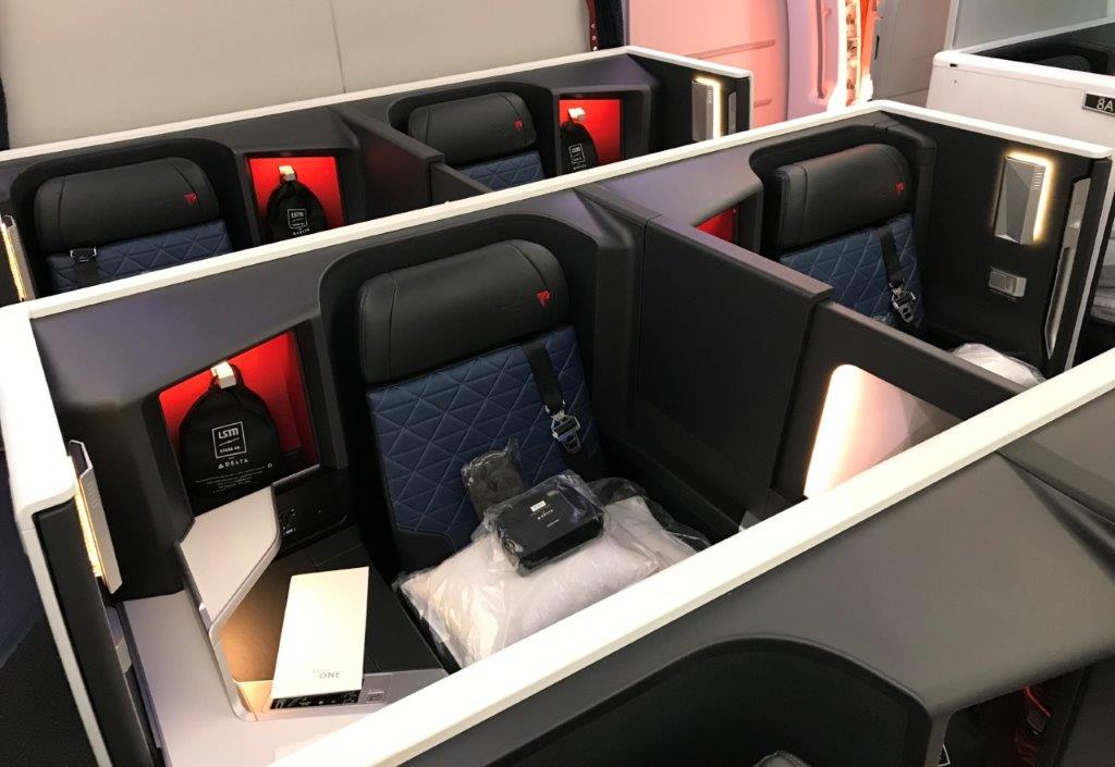 Four luxurious airline business class seats with privacy dividers, quilted navy and black upholstery, red accents, and amenities including pillows and packaged comfort kits placed on each seat.