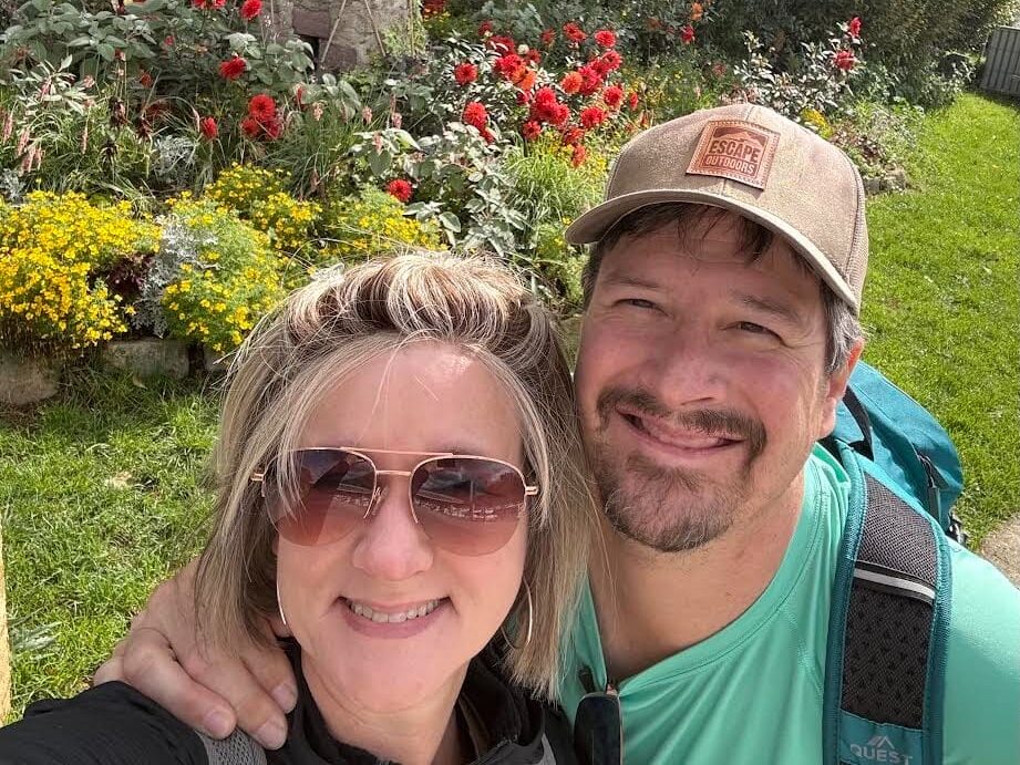 A couple stands smiling in front of a flower garden with a small windmill in the background. The woman wears sunglasses, and the man has a cap and a backpack. Bright red and yellow flowers surround them, and trees can be seen behind the windmill.