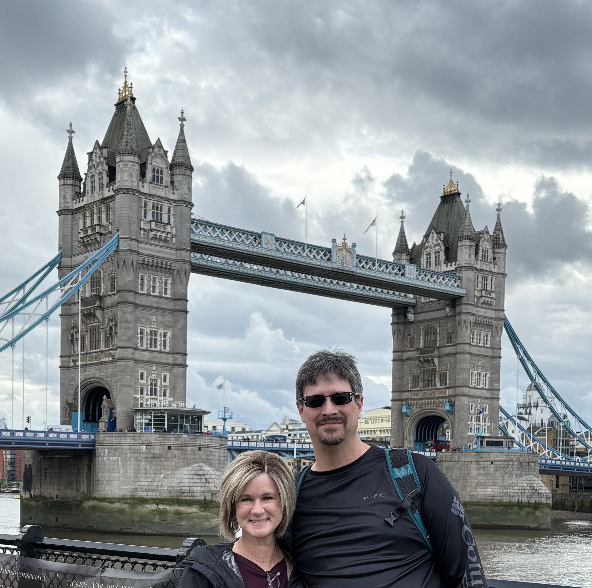A man and woman stand in front of Tower Bridge in London, smiling. The bridges iconic twin towers and suspension elements are visible against a cloudy sky. The Thames River flows below.
