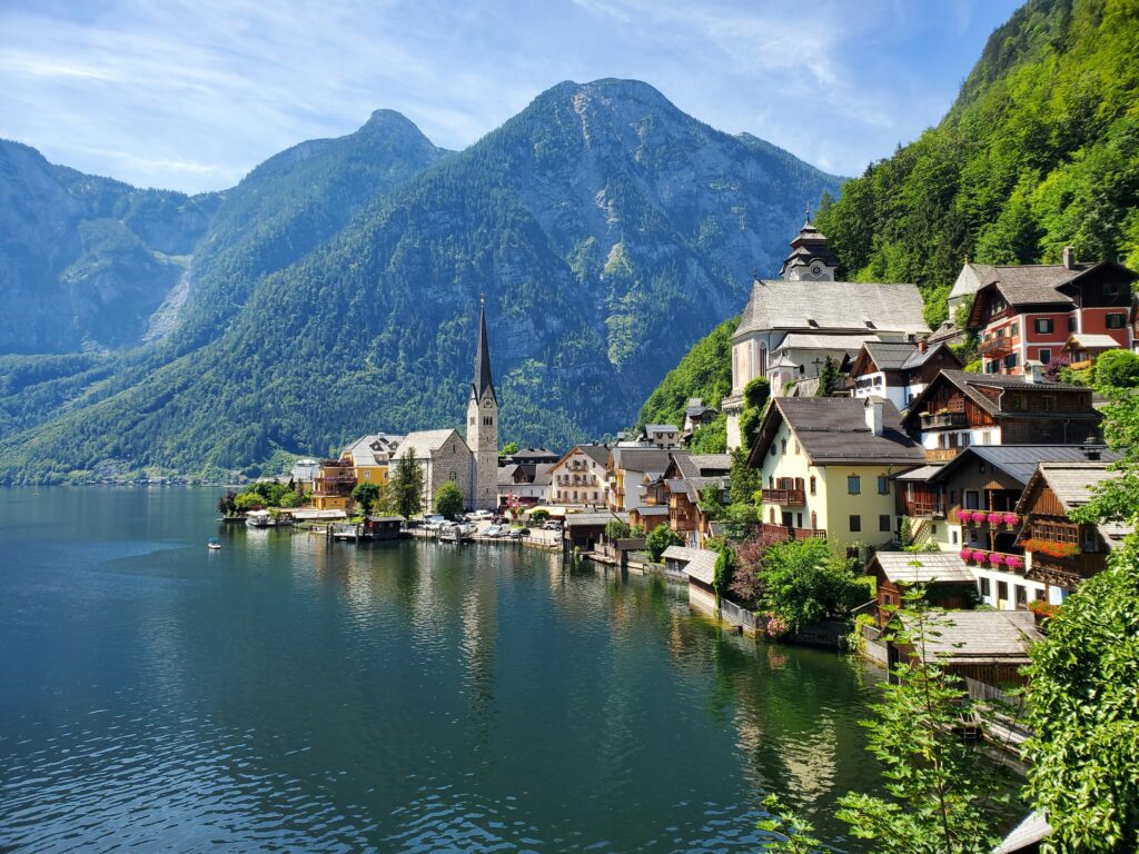 A picturesque lakeside village with colorful houses, a church steeple, and lush green mountains in the background, reflected in calm water under a clear blue sky.