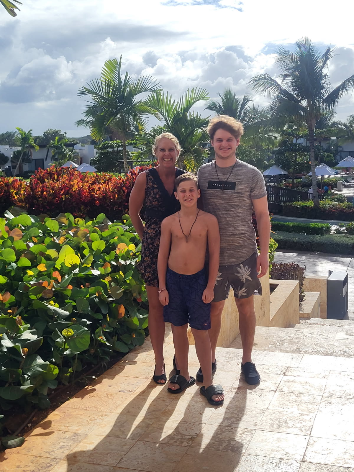 A woman and two boys stand smiling on stone steps surrounded by lush greenery and palm trees, with a pool and resort buildings in the background under a cloudy sky.