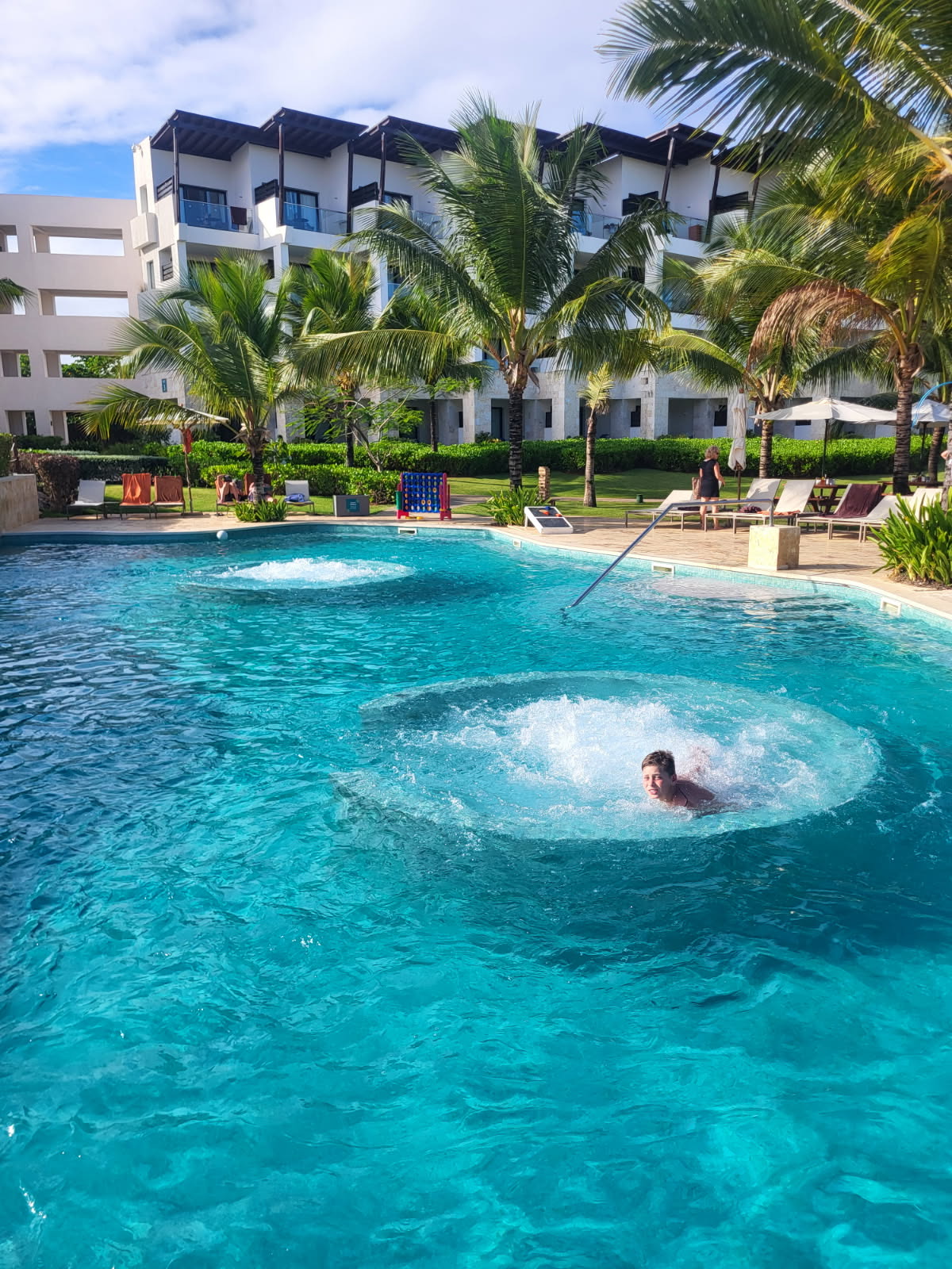 A person is swimming in a clear blue pool with bubbling water, surrounded by palm trees and a modern white hotel building in the background under a partly cloudy sky.