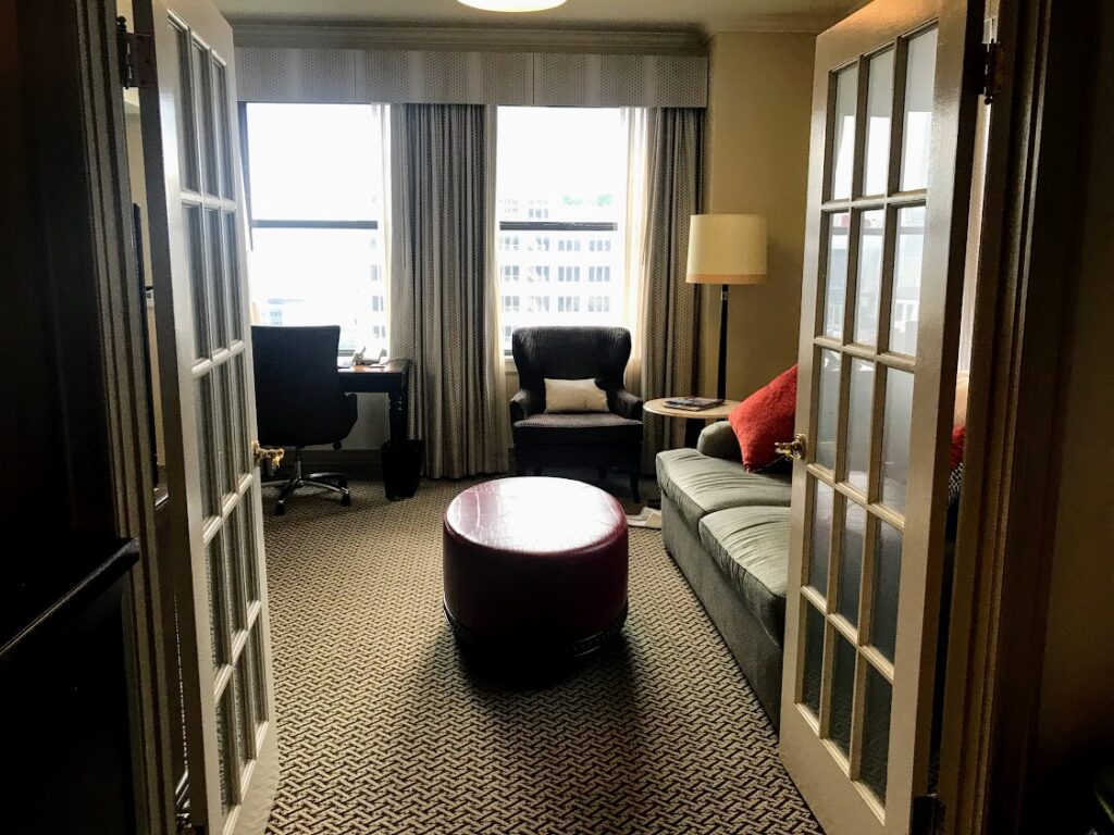 A cozy living room with French doors, a gray sofa with orange pillows, a purple ottoman, an armchair by the window, a floor lamp, and a desk with a chair near large windows letting in natural light.