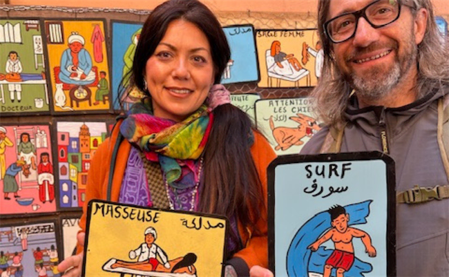 Two people smiling and holding colorful illustrated signs, one reading “Masseuse” and the other “Surf,” with more vibrant illustrated signs displayed on a wall behind them.