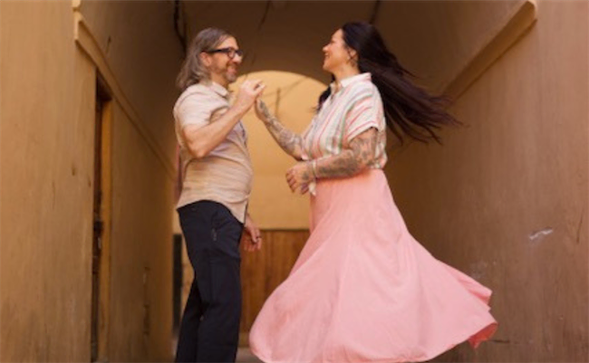 A man and woman smile and dance together in a narrow, sunlit hallway. The woman’s pink skirt swirls as she twirls, and both appear joyful and relaxed, enjoying the moment.