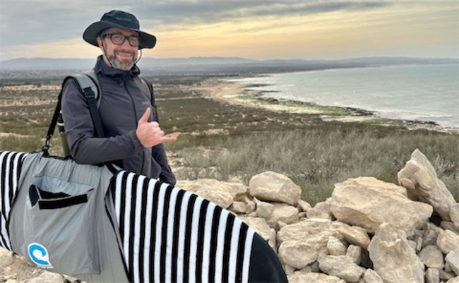 A man in a hat and glasses stands near a rocky shore, smiling and making a “shaka” hand gesture. He carries a striped surfboard bag with the ocean and a sandy coastline visible in the background.