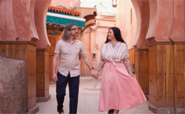 A couple holding hands, smiling at each other, walk under a decorative archway in a sunlit, warm-toned, outdoor setting with wooden and stone elements.