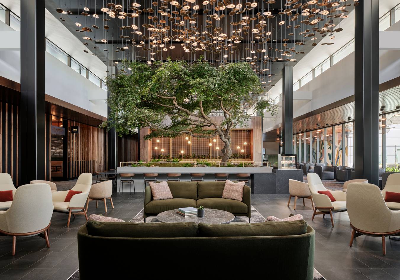 Modern, spacious hotel lobby with contemporary seating, a large tree as a centerpiece, pendant lights overhead, and floor-to-ceiling windows letting in natural light.