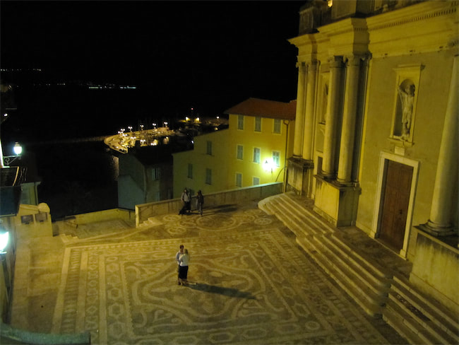 A person stands in the center of an ornate, patterned courtyard at night, near a large, illuminated building with steps. In the background, a few people and lit buildings overlook a dark waterfront.