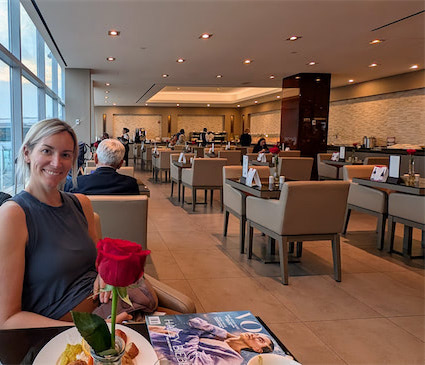 A woman sits at a table with a red rose, dessert, and a magazine in a spacious, modern lounge filled with tables and people dining. Large windows line one side of the room, letting in natural light.
