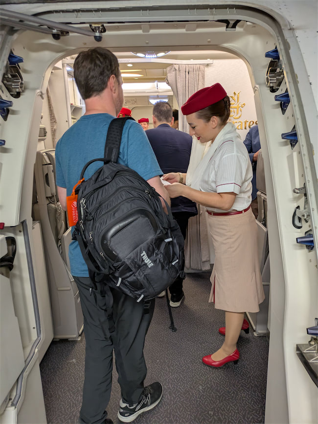 A man with a black backpack boards an Emirates airplane, handing his ticket to a flight attendant wearing a beige skirt, white shirt, and red hat near the aircraft entrance.
