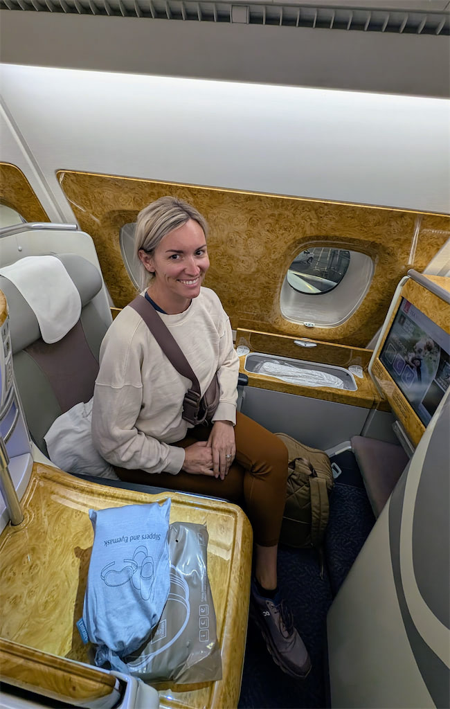 A woman with blonde hair, wearing a cream sweater and brown pants, sits in a luxurious first-class airplane seat near a window, smiling at the camera. A travel amenity kit is on the seat tray in front of her.