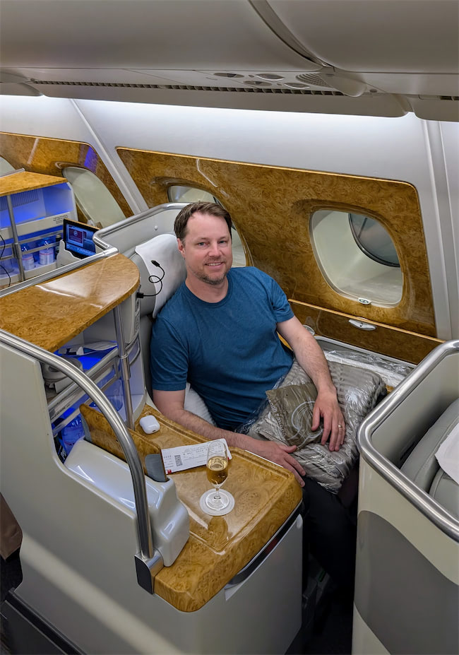 A man in a blue shirt smiles while seated in a luxurious first-class airplane seat, with wood paneling, a drink, a menu, and a blanket, next to large windows.