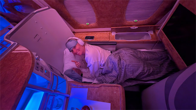 A person lies comfortably under a blanket in a luxurious airplane first-class suite, wearing headphones and a sleep mask on their head, smiling and giving a thumbs up. The cabin is lit with soft blue and purple lighting.