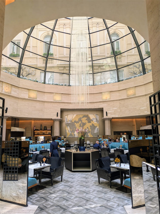 A luxurious restaurant with a glass-domed ceiling, elegant seating, and a large abstract artwork on the back wall. Natural light fills the space, and diners are seated at tables with blue accents. Staff are serving guests.