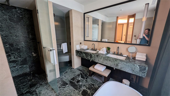Modern hotel bathroom with green marble floor and counters, double sink, large mirror, glass shower, bathtub, and a person partially visible in the reflection, holding a phone. Towels and toiletries are neatly arranged.