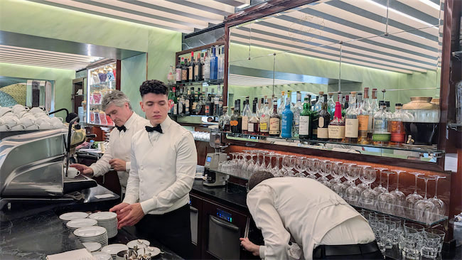 Three bartenders in white shirts and black bow ties work behind a bar lined with glasses, bottles of liquor, and an espresso machine under a striped ceiling.