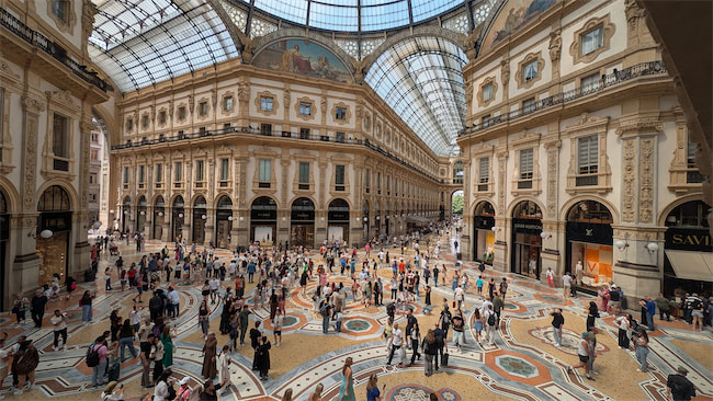 A large crowd of people walks and gathers inside a grand, glass-roofed shopping arcade with ornate architecture and colorful mosaic floors. Sunlight streams through the vaulted glass ceiling.