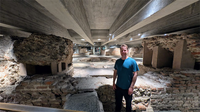 A man in a blue shirt stands among ancient stone and brick ruins beneath a modern concrete ceiling with spotlights, creating a blend of historic and contemporary architecture.
