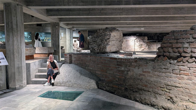 A woman sits on a bench inside an underground archaeological site with exposed ancient brick walls and ruins. In the background, two people look at displays and artifacts under concrete ceilings.