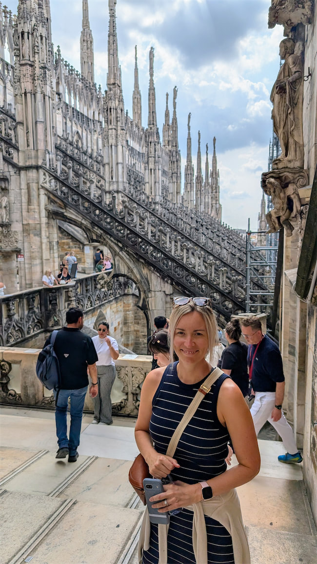 A woman in a striped dress smiles at the camera while standing on the rooftop of the Milan Cathedral, surrounded by intricate Gothic spires and other visitors exploring the landmark.