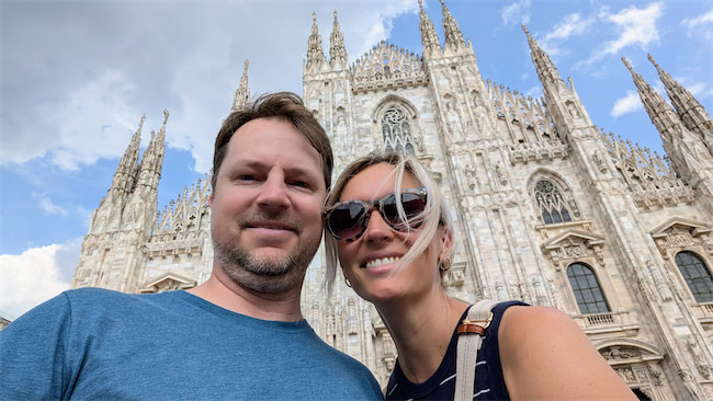 A man and woman smiling in front of the ornate facade of the Milan Cathedral (Duomo di Milano) on a partly cloudy day. The cathedrals intricate architecture is visible in the background.