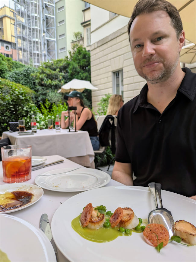 A man sits at an outdoor restaurant table with a plate of scallops on green puree, a cocktail, and other diners in the background, set against a cityscape and greenery.