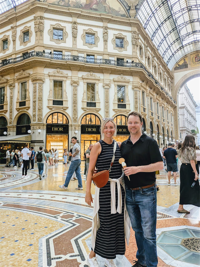 A smiling couple poses inside a grand, ornate shopping arcade with mosaic floors and a glass-domed ceiling. Behind them are Dior storefronts and several people walking around.