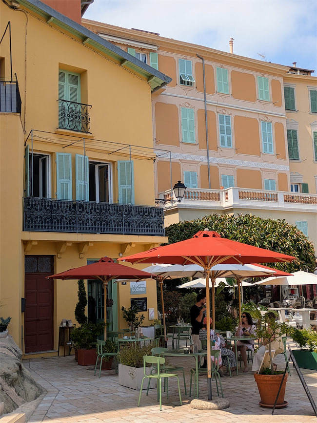 Outdoor café with red umbrellas, green chairs, and potted plants in front of pastel-colored buildings with shutters and balconies on a sunny day. People are sitting and relaxing at the tables.