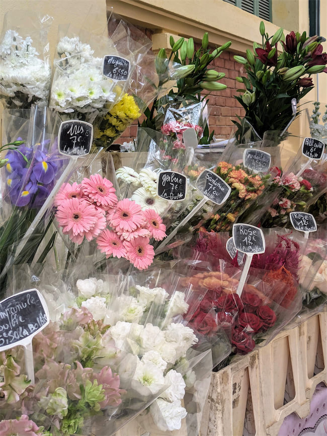 Bouquets of colorful flowers wrapped in plastic are displayed for sale outdoors, each bunch labeled with handwritten price tags. The flowers include roses, lilies, gerberas, and other assorted blooms.