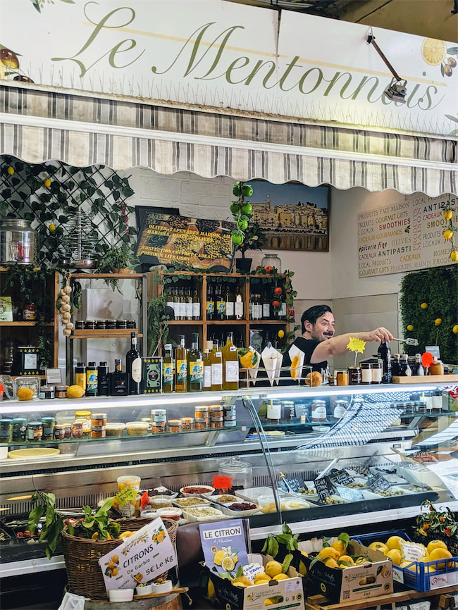 A cheerful vendor stands behind a shop counter decorated with lemons, bottles, and jars. The shop, named Le Mentonnais, displays fresh lemons, products, and signs in French. The ambiance is bright and inviting.