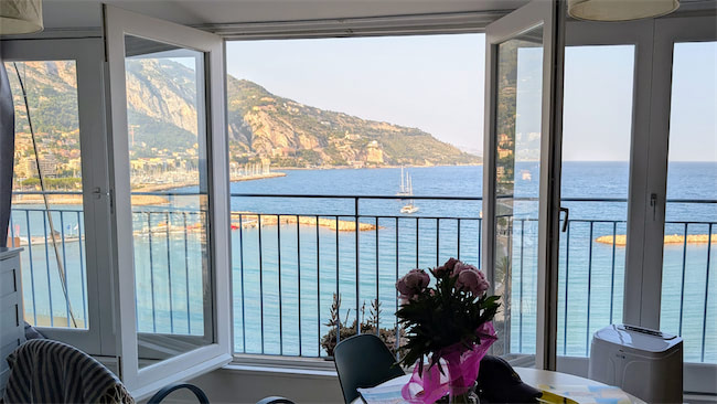 Large windows open to a scenic view of the sea, mountains, and sailboats. A vase of pink flowers sits on a table inside, with natural light brightening the airy, inviting room.