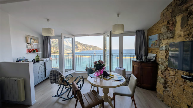 A bright, modern living room with large open windows overlooking a blue sea and mountains. Theres a round table with flowers, chairs, a rocking chair, and shelves and cabinets along the walls. Sunlight fills the space.