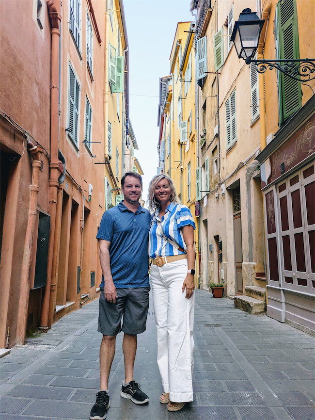 A man and a woman stand smiling in a narrow, colorful European street lined with tall, orange and yellow buildings with green shutters. The street is paved with stone tiles and lit by a wall-mounted lamp.