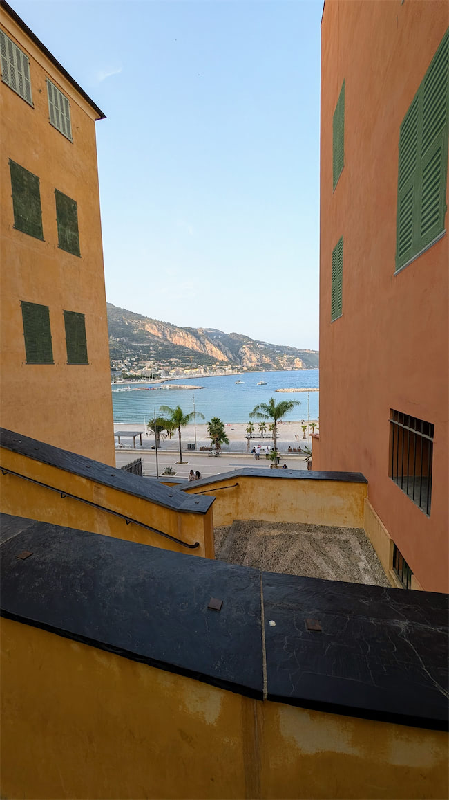 View between two tall, colorful buildings with green shutters, looking toward a beach, palm trees, and mountains in the distance under a clear blue sky. A zigzag ramp leads down to the promenade.