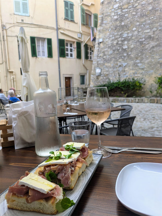 A wooden table set with a plate of bruschetta topped with prosciutto and cheese, a glass and bottle of water, a glass of rosé wine, and a white plate, in an outdoor cafe beside old stone buildings.