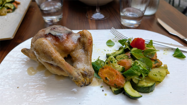 A roasted small bird, possibly a Cornish hen, is served on a white plate with a side of mixed sautéed vegetables including carrots, zucchini, and greens. Glasses of water and utensils are in the background.