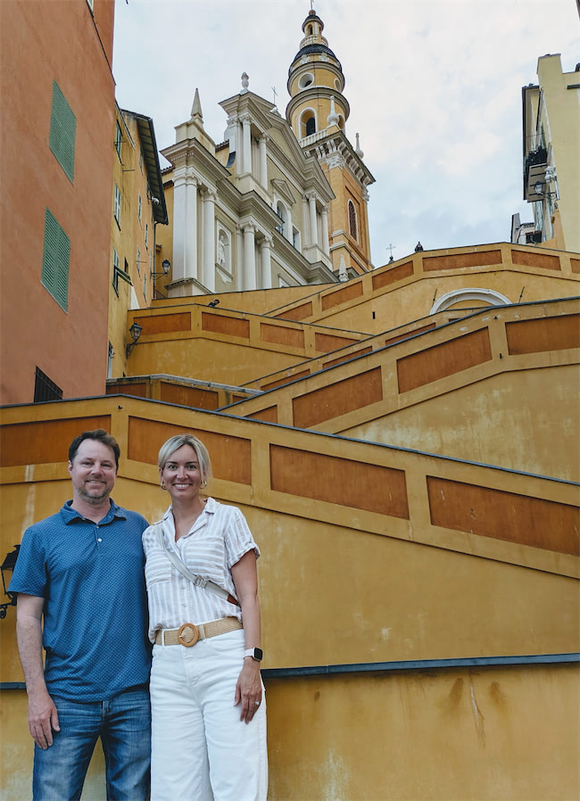 A man and woman stand smiling in front of wide, yellow-orange terraced steps leading up to a historic church with a tall bell tower in a European town. The sky is light blue with some clouds.