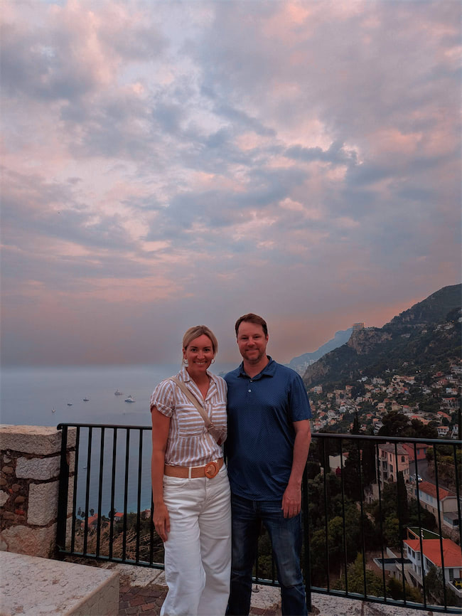 A woman and man stand together in front of a black railing overlooking a coastal town with mountains and the sea in the background, under a pastel-colored, cloudy sky at sunset.
