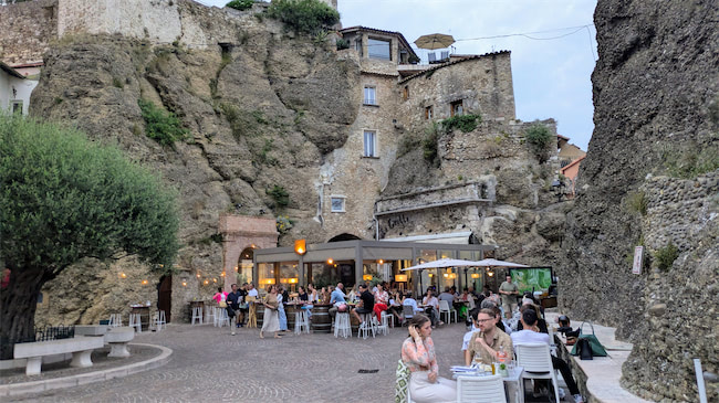 People dine at outdoor tables in a lively restaurant set against rocky cliffs and old stone buildings, with greenery and evening lights creating a cozy atmosphere.