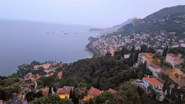 A coastal town with red-roofed houses sprawls along a lush, tree-covered hillside overlooking the calm blue sea, with boats anchored offshore and mountains in the distance under a hazy sky.