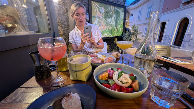 A woman sits at an outdoor restaurant table with plates of colorful salad, bread, drinks, and a map display behind her. She looks at her phone while the street and buildings are visible in the background.