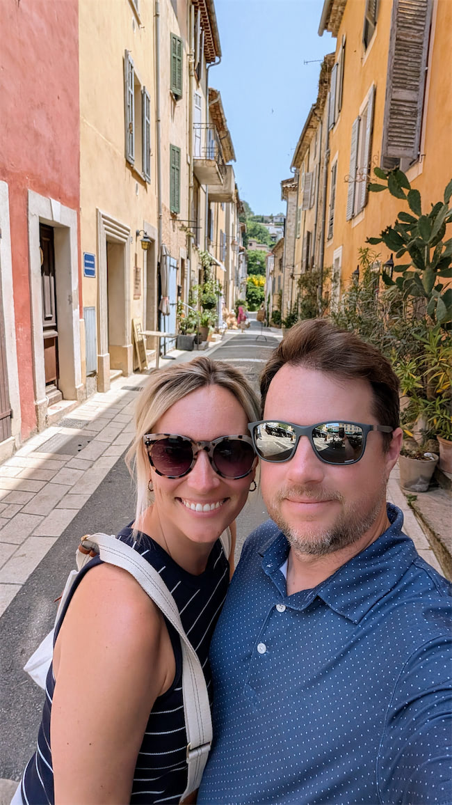 A smiling couple wearing sunglasses takes a selfie on a sunny, narrow street lined with colorful buildings and plants. The scene suggests a charming European town.